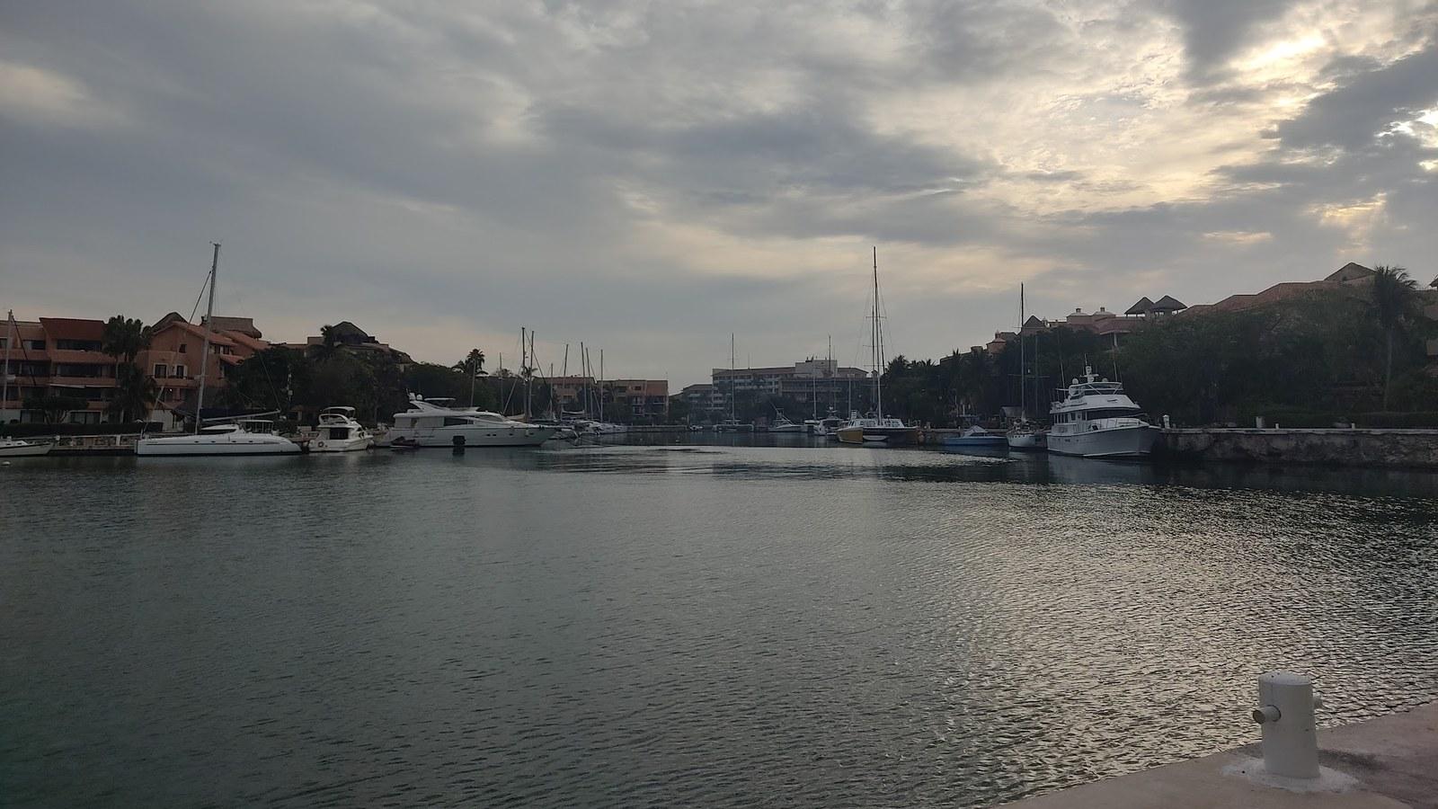 Marina de Puerto Aventuras bajo un cielo nublado, con yates y veleros atracados, casas con frente de agua al fondo y un muelle de piedra en primer plano