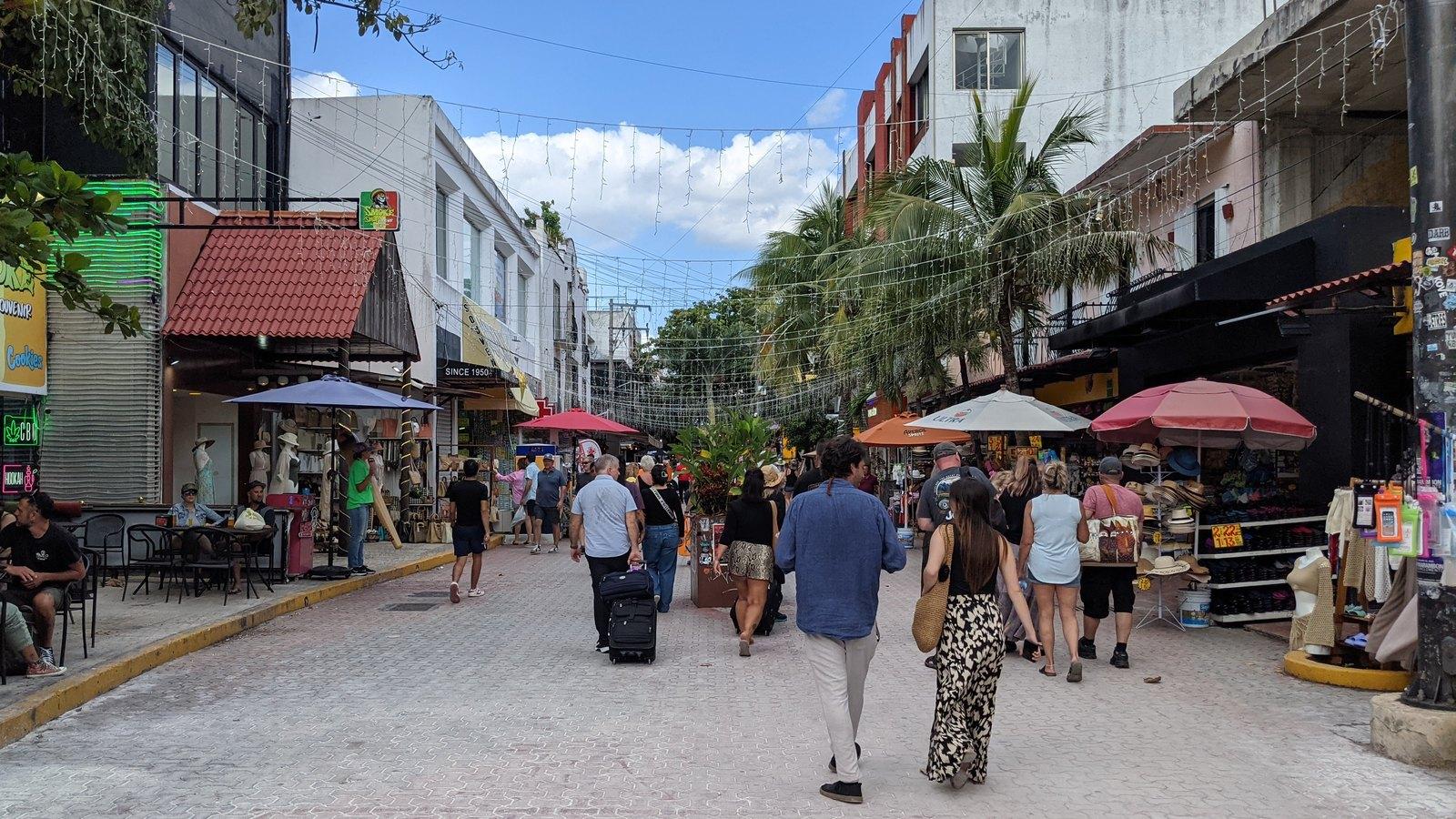 La Quinta Avenida de Playa del Carmen en un día soleado, con gente caminando por la calle peatonal entre comercios, sombrillas y palmeras